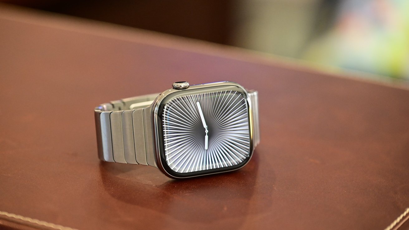 A sleek smartwatch with a silver metal band sits on a brown surface, displaying a radial black-and-white pattern on its screen.