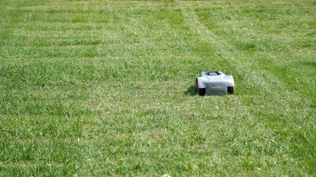 Robotic lawnmower operating on a large, evenly-cut grassy field under a clear sky.
