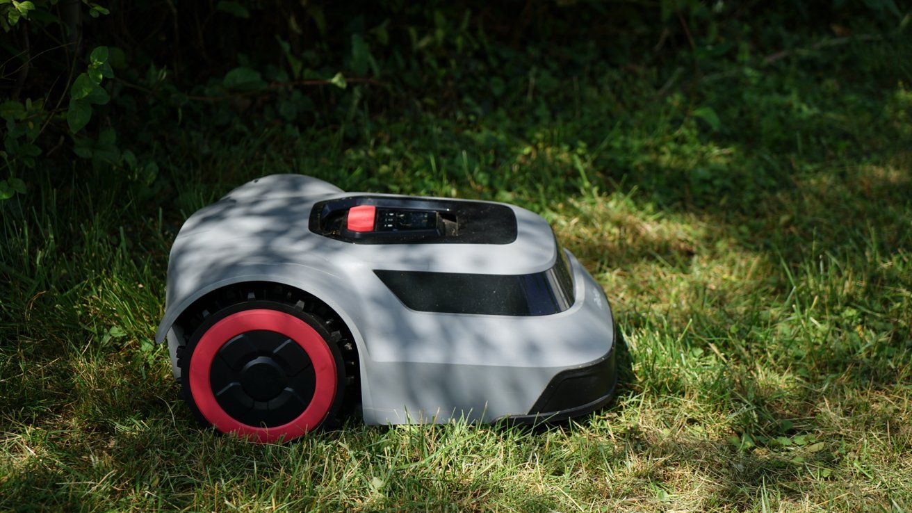 Robotic lawn mower on grass with a red and gray body, partially shaded by trees.
