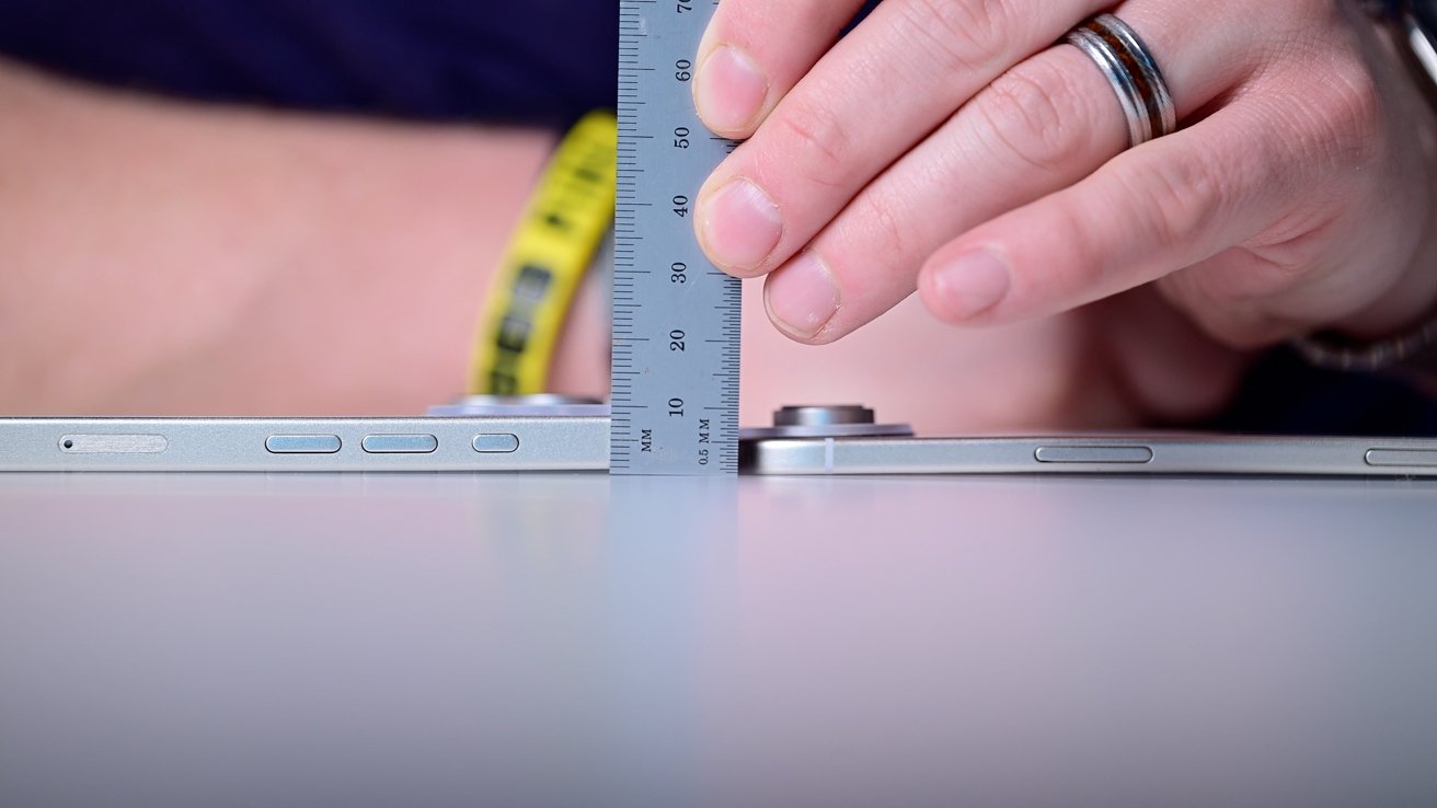 Hand measuring thickness of two smartphones with a ruler on a table. Both devices have visible side buttons and protruding cameras.