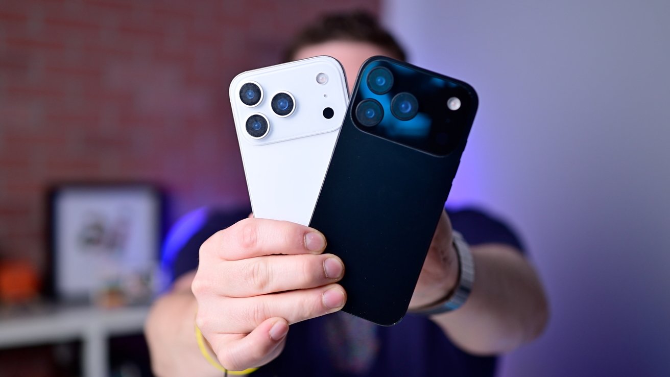 A person holds two smartphones, one white and one black, both showcasing triple camera setups on the back.