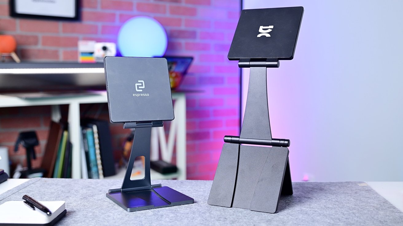 Two black tablet stands on a gray surface with a blurred background featuring a brick wall and shelves.