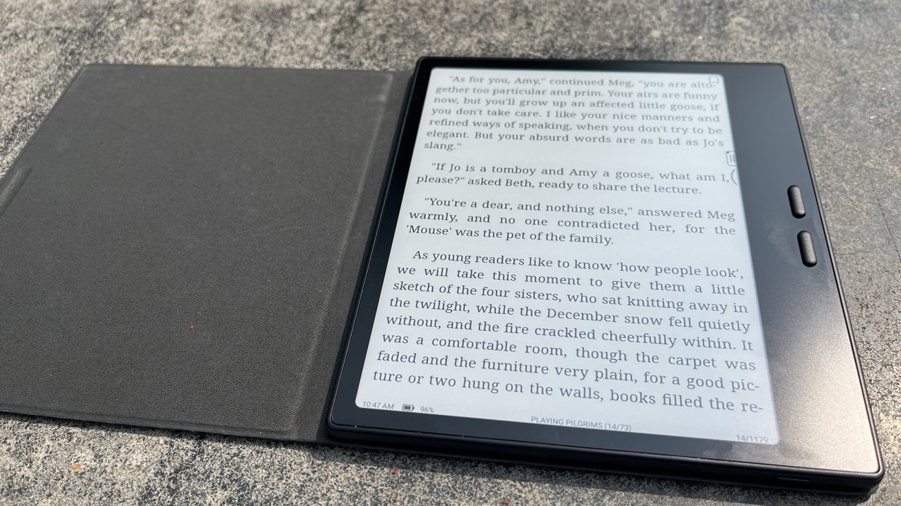 E-reader displaying text from a book, placed on a surface with its protective cover open. The screen shows a page from 'Playing Pilgrims' with reading progress indicated.