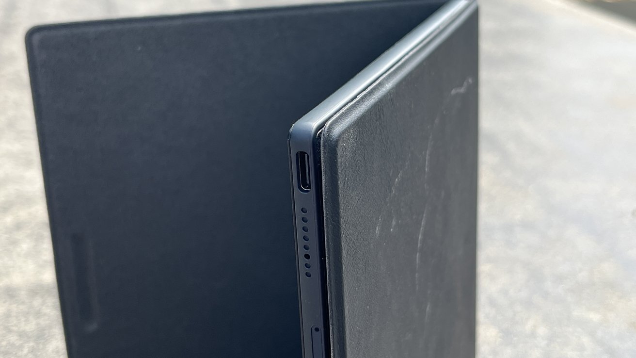 Close-up of a thin, dark-colored, folding electronic device with a visible USB-C port and speaker holes along the edge, resting on a light surface.