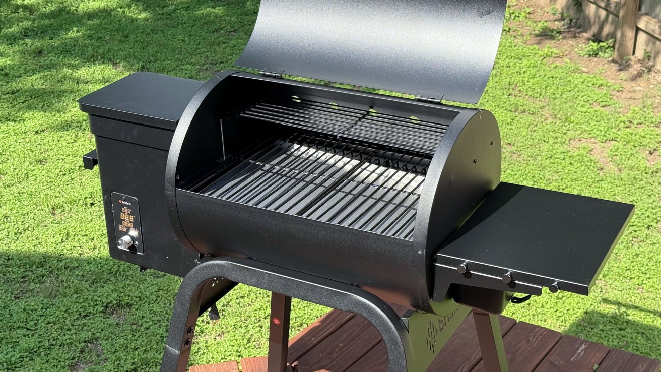 Outdoor black pellet grill with lid open, exposing grates. Electronic control panel and side shelf visible, set on wooden deck with grassy background.