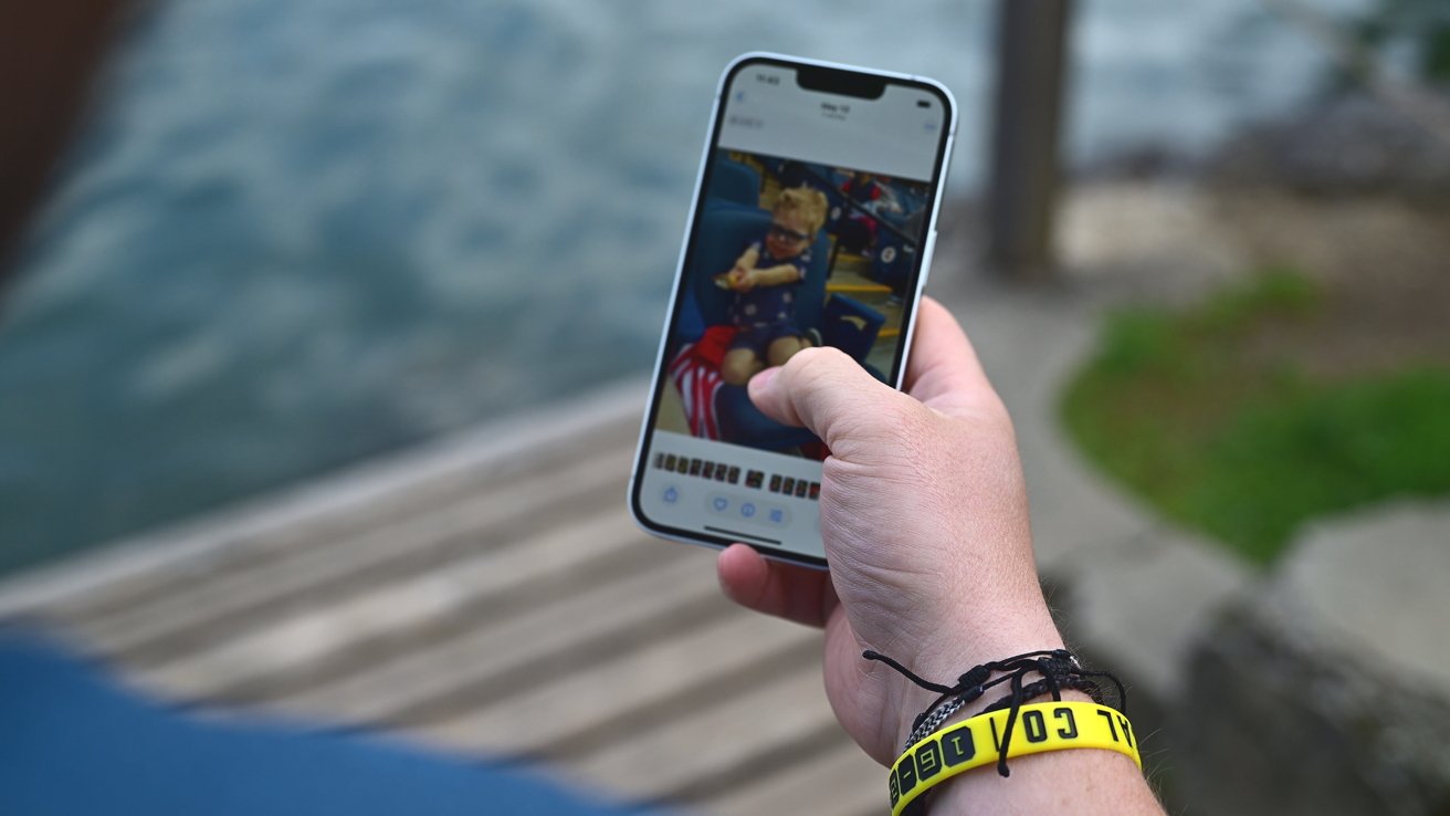 Hand holding a smartphone displaying a photo of a child in sunglasses. Blue water and wooden deck visible in the background. Yellow wristband on wrist.