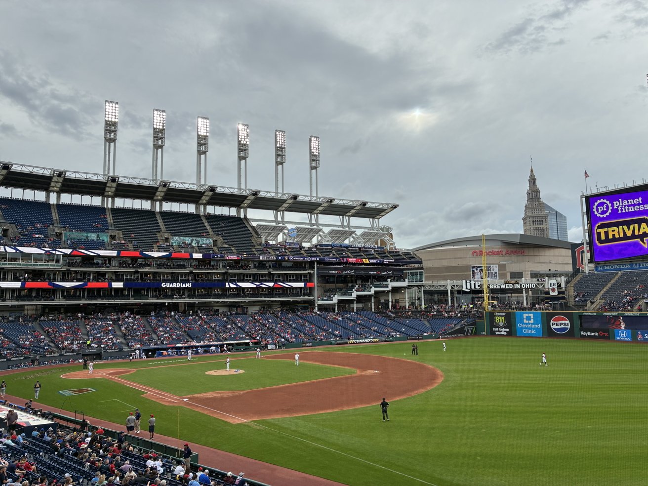 Baseball stadium with players on the field, spectators in the stands, large digital screen displaying advertisements, and a cityscape with tall buildings in the background under a cloudy sky.