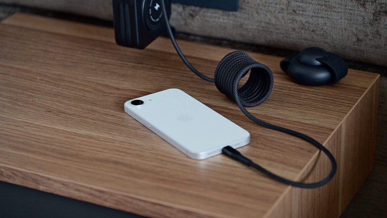 White smartphone charging with a black braided cable on a wooden surface, next to a coiled cable and a small black object.