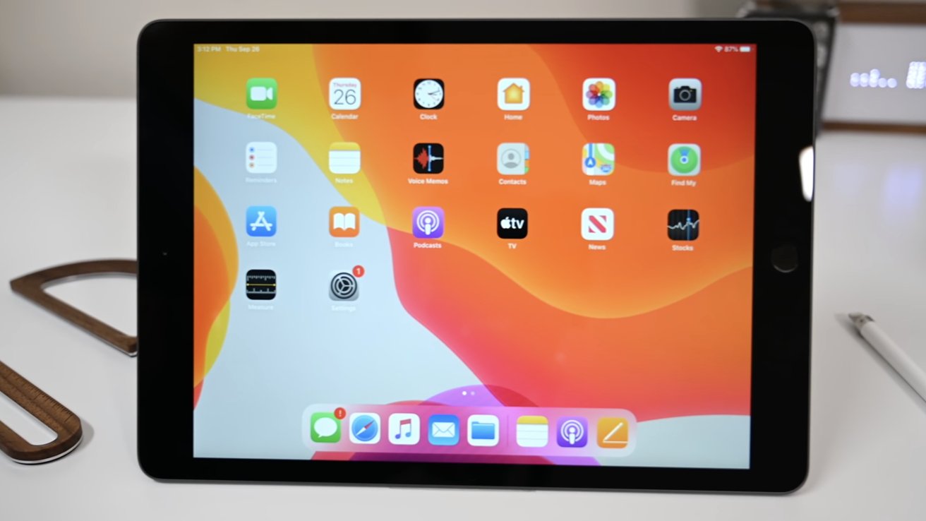 Tablet displaying various app icons on a screen with an orange and yellow wavy background, placed on a white surface with a stylus nearby.